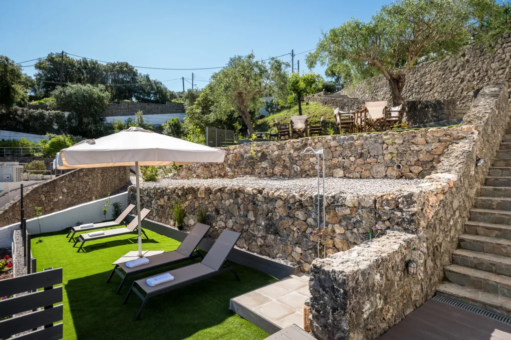 Stone walled terrace with sun loungers and a large umbrella over artificial grass overlooking a seating area on a higher level surrounded by trees