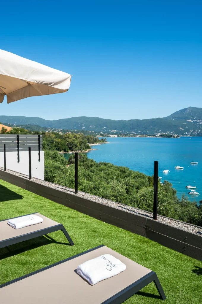 Sun loungers with towels on a grassy terrace overlooking a blue lake and distant mountains under a clear sky