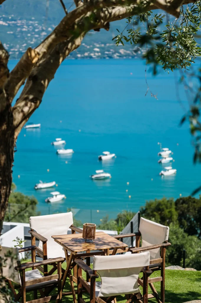 Wooden outdoor dining table and chairs on a grassy terrace overlooking a blue bay with small boats floating nearby