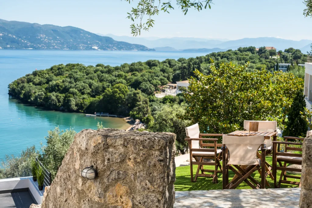 Outdoor terrace with a wooden dining set overlooking a turquoise bay lush greenery and distant hills A rocky wall sits in the foreground