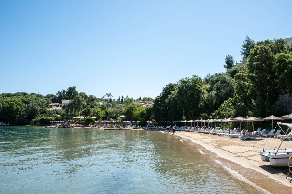 Beach scene with rows of sun loungers and thatched umbrellas along a tree lined shore and calm blue water