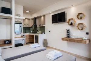 Modern hotel suite with two twin beds, a kitchenette, and wall-mounted TV; towels on beds ready for guests.