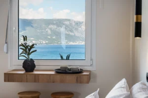Window with a seaside view: blue sea and distant mountains seen through a white frame; a potted plant and two teacups on a wooden shelf below.