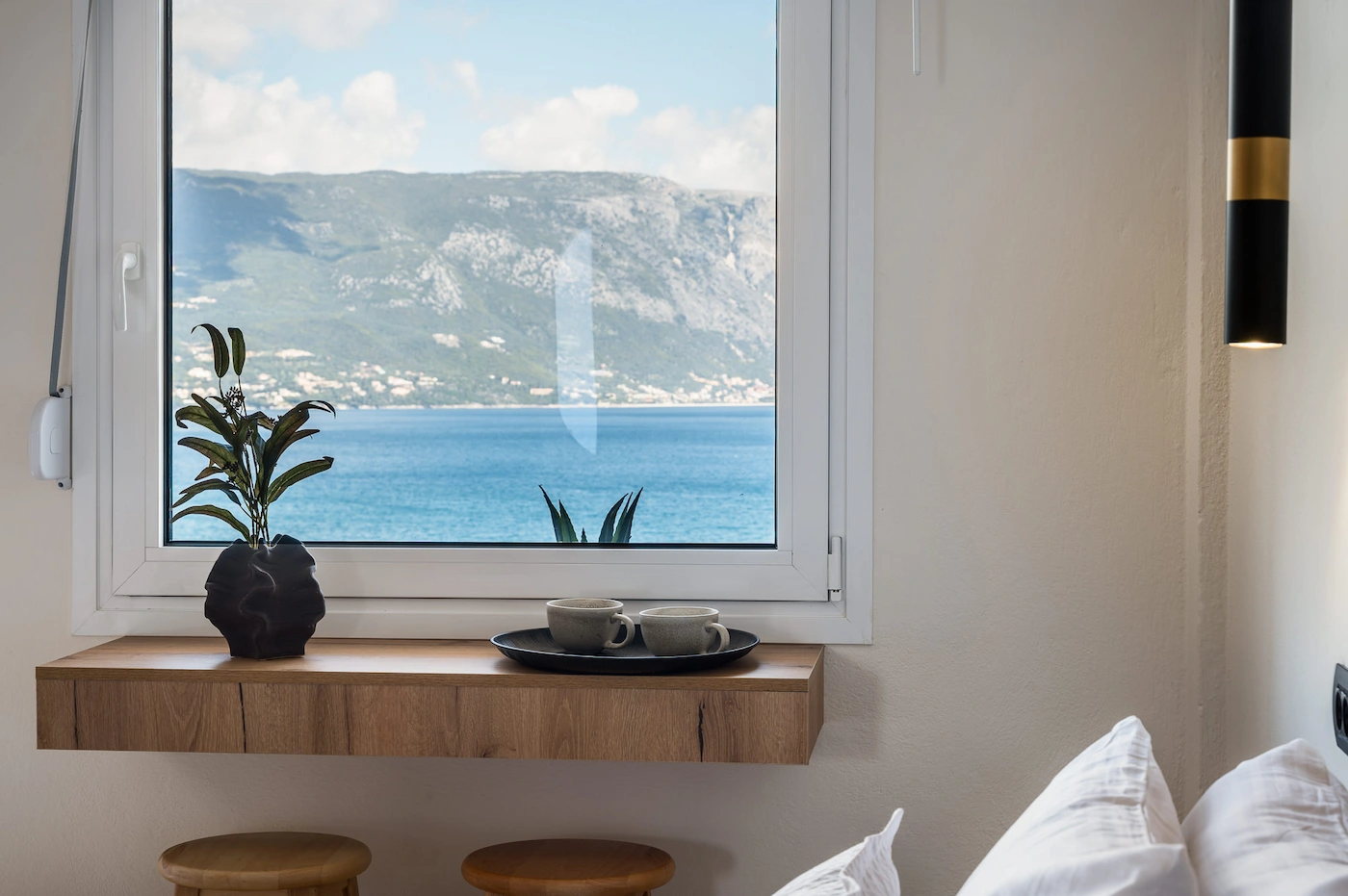 Window with a seaside view: blue sea and distant mountains seen through a white frame; a potted plant and two teacups on a wooden shelf below.