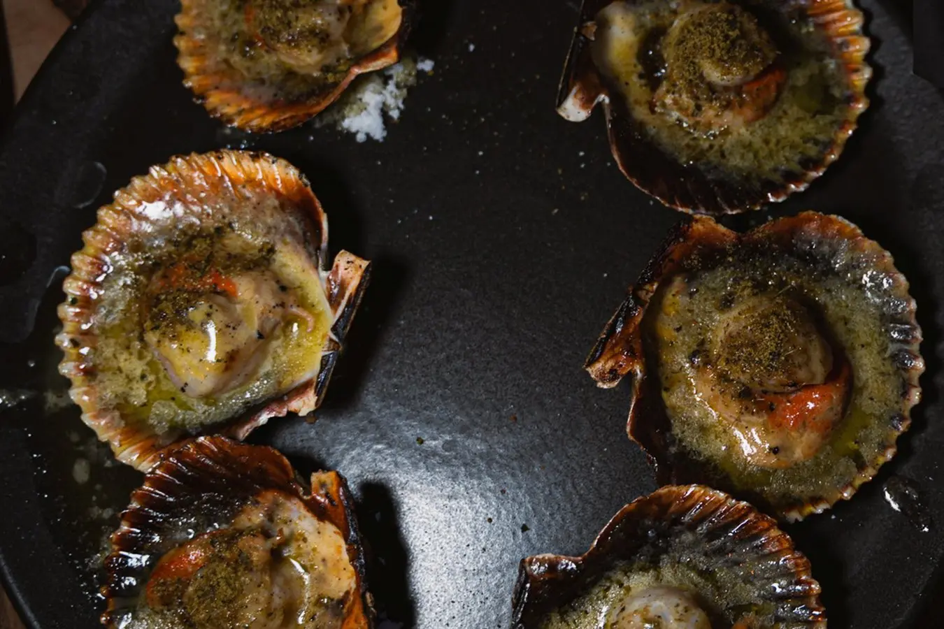 Six baked scallops in their shells arranged in a skillet, topped with herbs and melted butter.