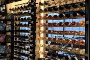 Wine bottles arranged on wooden racks in a shop display, organized by color with warm lighting highlighting the rows of red, white, and rosé bottles.