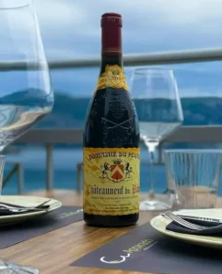 Bottle of red wine on a dining table set for a meal, with empty wine glasses and a sea‑facing balcony in the background.