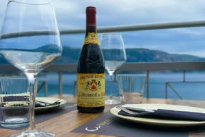 Bottle of wine on a dining table with crystal glassware, overlooking a sea view and mountains in the background.